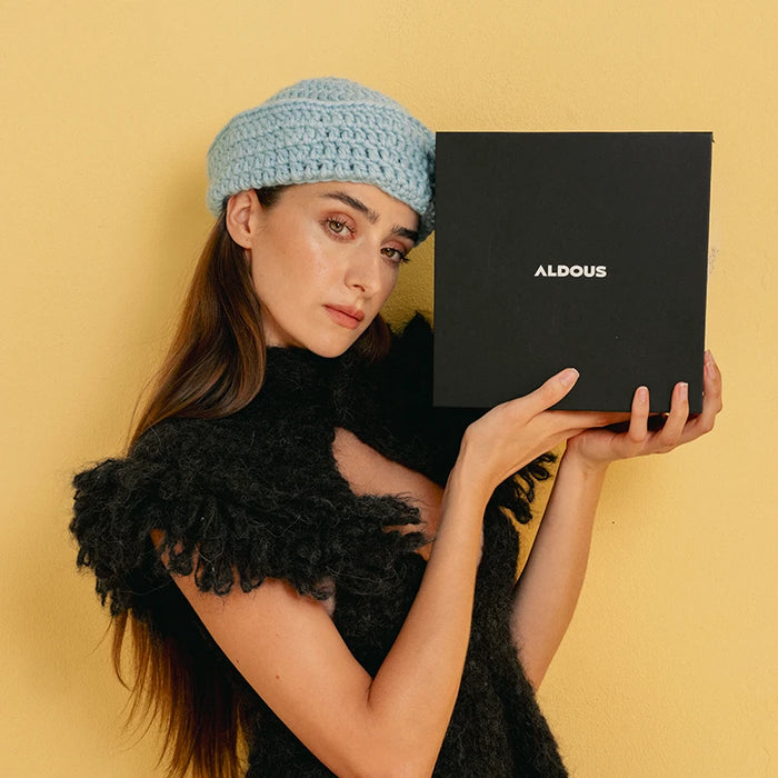 Modelo con gorro de punto azul claro y chal negro texturizado, sosteniendo una caja de regalo negra con el logo de Aldous.